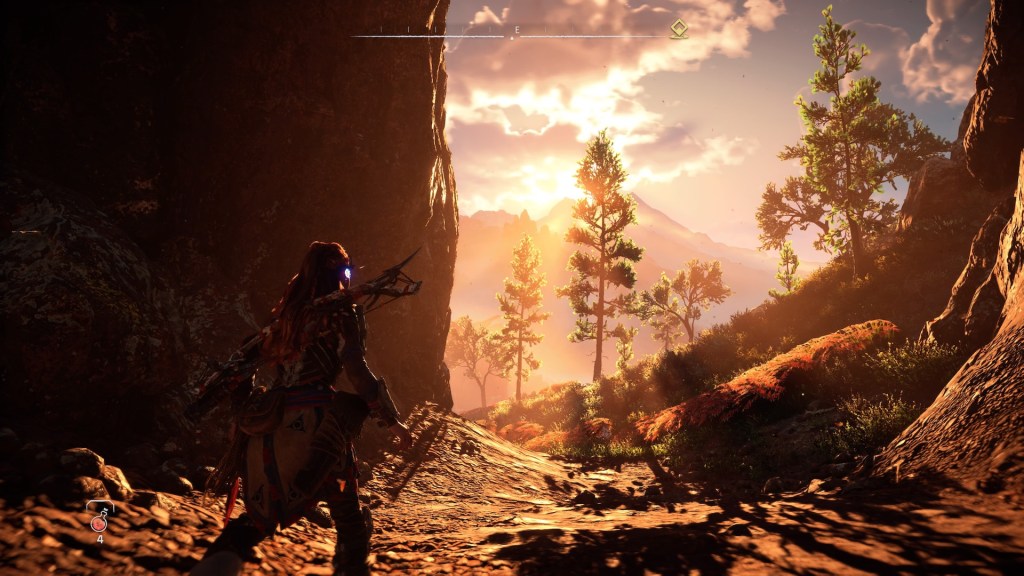Aloy standing in a cave looking at a colorful sundown behind a ridge of mountains.