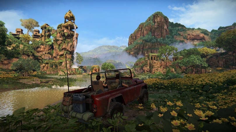 Chloe and Nadine in a red jeep in a field with water areas, yellow flowers, and mountains with ancient edifices in the distance in front of a blue sky.