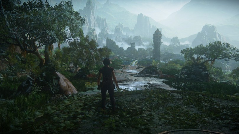 Chloe stands in the middle of a jungle-like area in the rain. Cloudy mountains can be seen in the distance. The road ahead of her is muddy.
